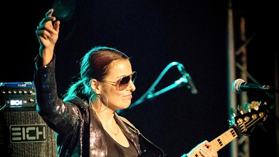 Die Musikerin Ida Nielsen steht mi einem Muskclub. Sie hat eine schwarze Bassgitarre in der Hand, die Haare sind zu einem Zopf nach hinten gekämmt, sie trägt eine Sonnenbrille und schaut ins Publikum.