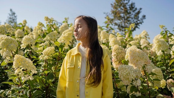Ein Kind steht mit geschlossenen Augen zwischen Blumen, die fast so groß sind wie das Kind