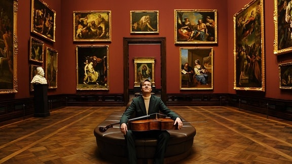 Jan Vogler sitzt mit einem Cello auf dem Schoß in der Dresdner Gemäldegalerie und schaut nach oben.