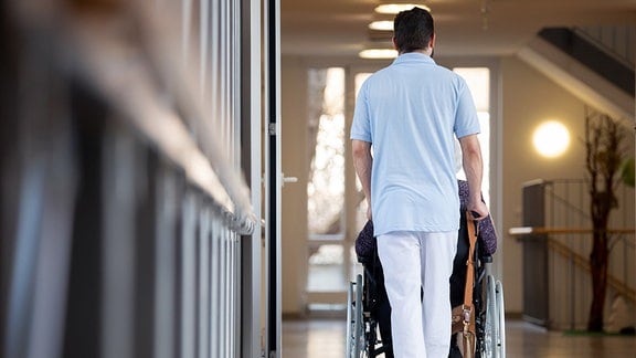 Ein Pfleger schiebt eine Bewohnerin eines Pflegeheims in einem Rollstuhl über den Flur.