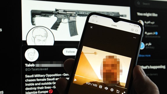 Smartphone zeigt das Gesicht eines Mannes, ein Laptop im Hintergrund eine Waffe