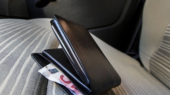 Symbolfoto: Einbruch ins Auto - Ein Dieb greift nach einer Brieftasche mit herausschauendem Geldschen, die in einem Auto liegt.