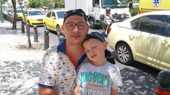 ein Vater hockt neben seinem kleinen Sohn. Die beiden blicken in die Handykamera der Mutter, die beide fotografiert.