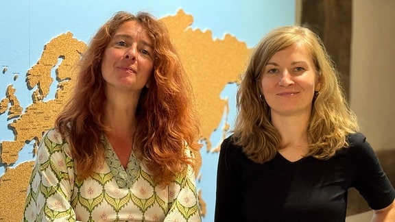 Zwei Frauen mit langen Haaren posieren leger für ein Foto vor einer Karte von Europa und Asien.