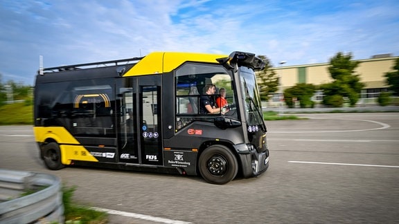 Moderner, kleinerer Bus in schwarz, mit gelben Farbakzenten, fährt auf Straße, Personen sitzen drin.