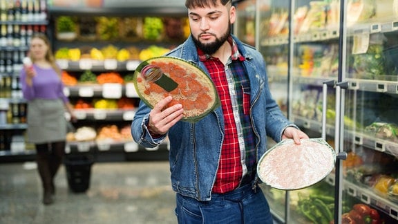 Mann steht im Supermarkt vor Kühlregalen, hält zwei eingeschweiste Tiefkühl-Pizzen in der Hand, schaut lesend auf hochgehaltene Salami-Pizza. Im Hintergrund unscharf Frau beim Einkauf.